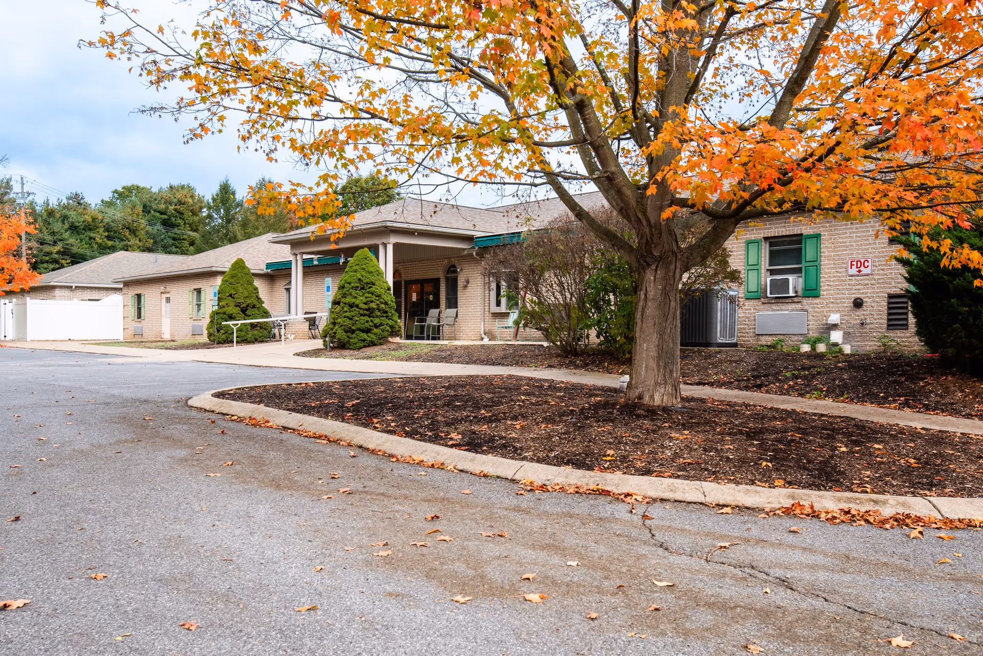 Exterior view of Salem Hill Haven Inc building with a paved driveway, landscaped area with a large tree with orange autumn leaves, green shrubs, and a covered entrance with chairs.