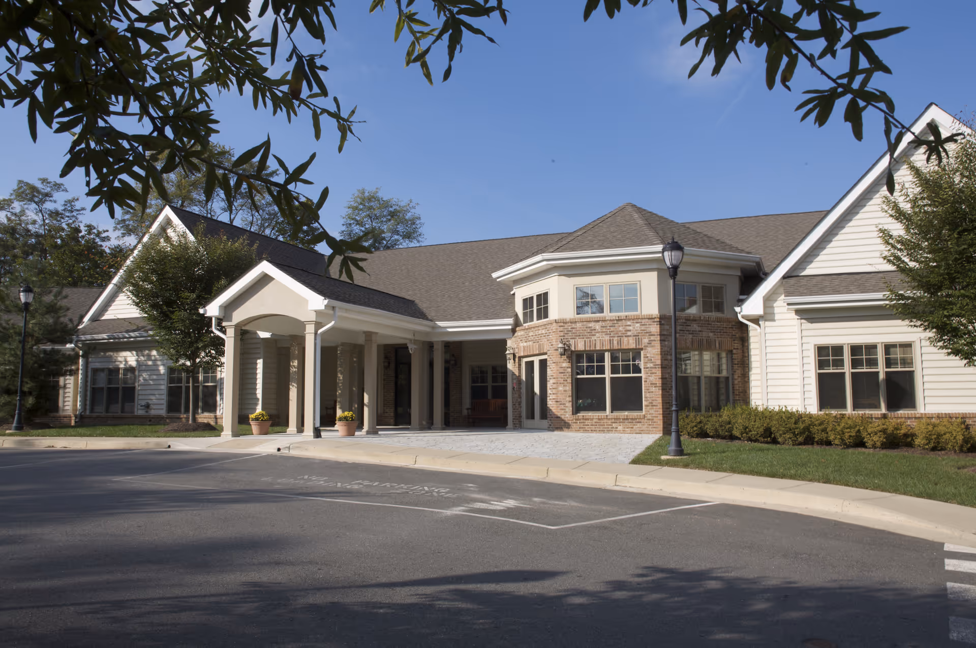 Exterior view of a single-story building with a covered entrance, large windows, and a combination of brick and siding walls, surrounded by trees and landscaping under a clear blue sky.