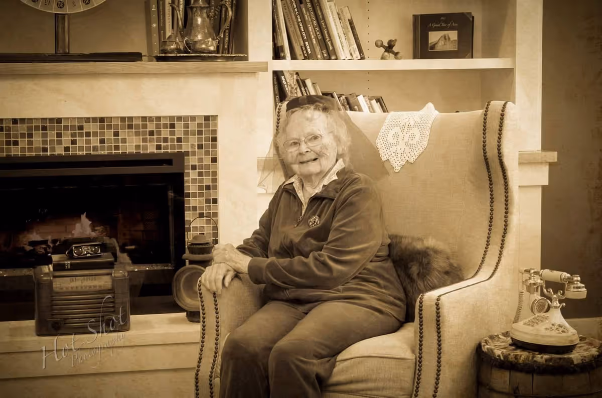 An elderly woman with glasses and a veil sits smiling in a cushioned armchair next to a fireplace with a lit fire. Behind her is a bookshelf with books and decorative items. A vintage rotary telephone is on a small round table beside the chair. The image has a sepia tone.