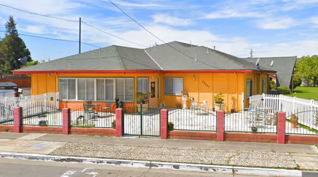 Single-story residential building painted in orange with a gray roof, surrounded by a white picket fence with red brick pillars. The front yard has a concrete patio with outdoor furniture and potted plants. The sky is partly cloudy.