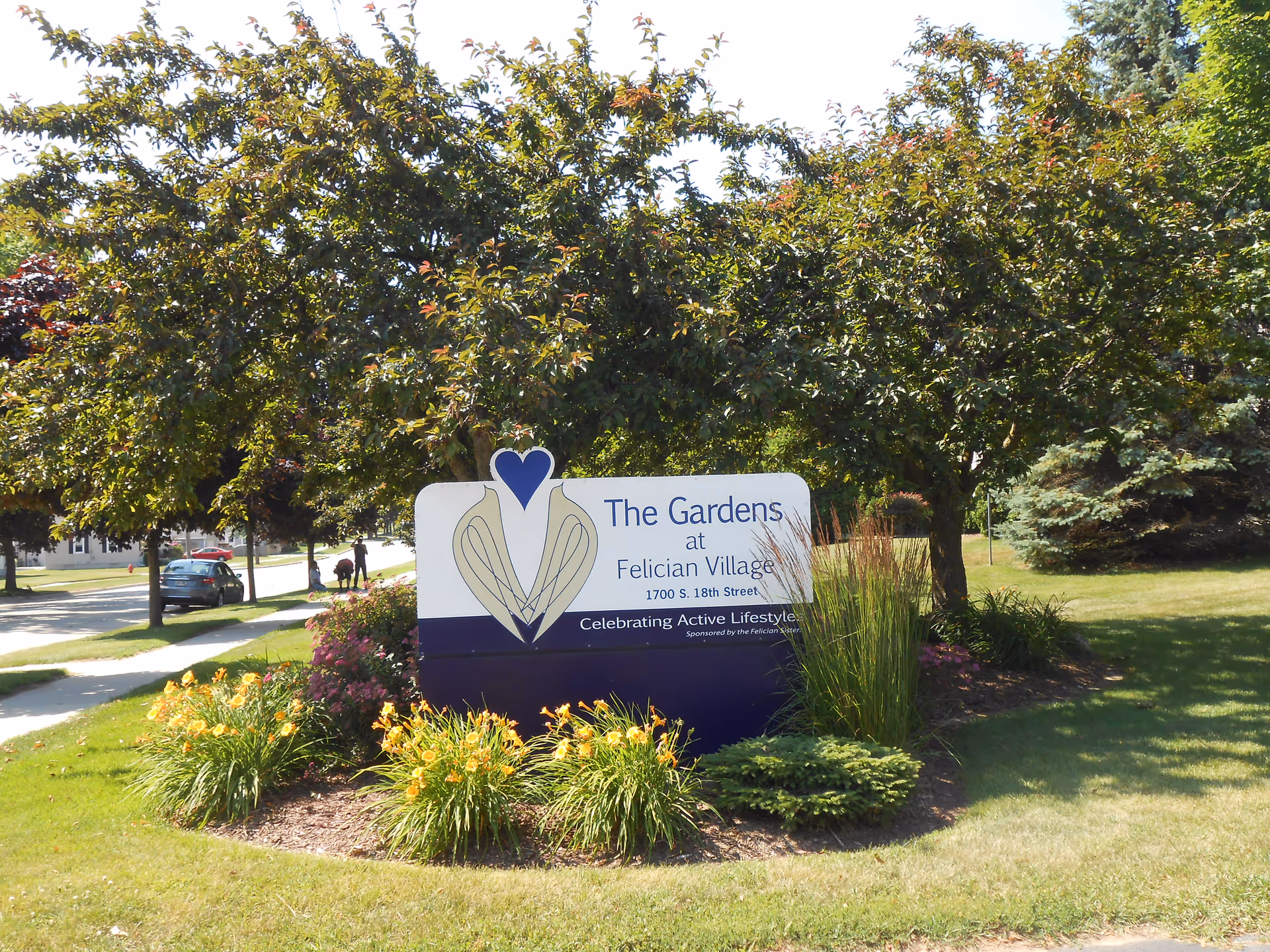 Outdoor sign for The Gardens at Felician Village, surrounded by trees and flowers.