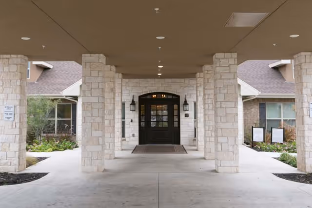 Covered porte-cochere with stone columns leading to a double-door main entrance of a senior living facility.