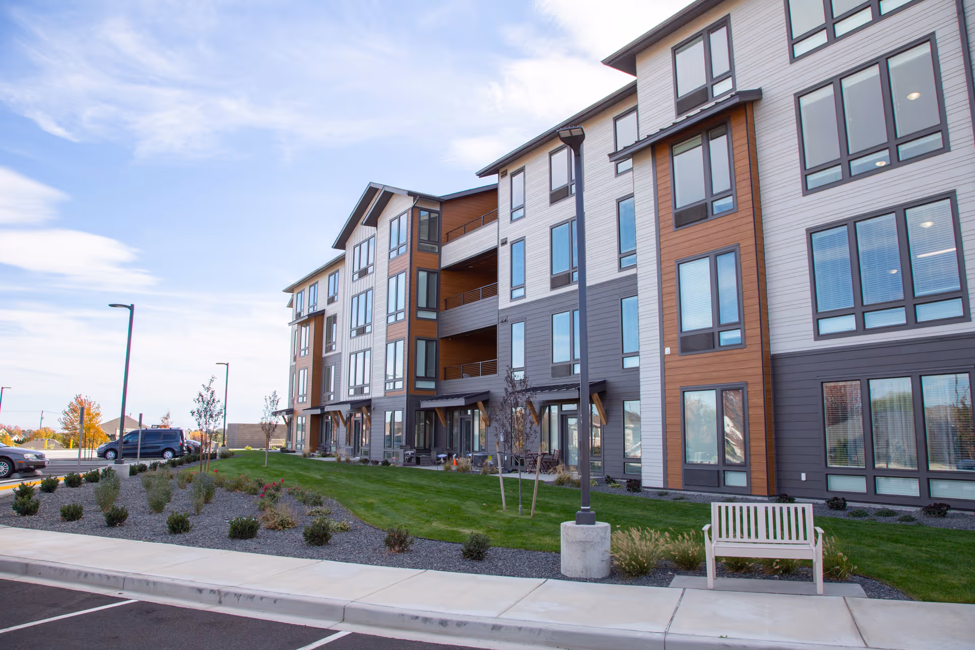 Modern multi-story senior living building with balconies, landscaped grounds, and a bench.