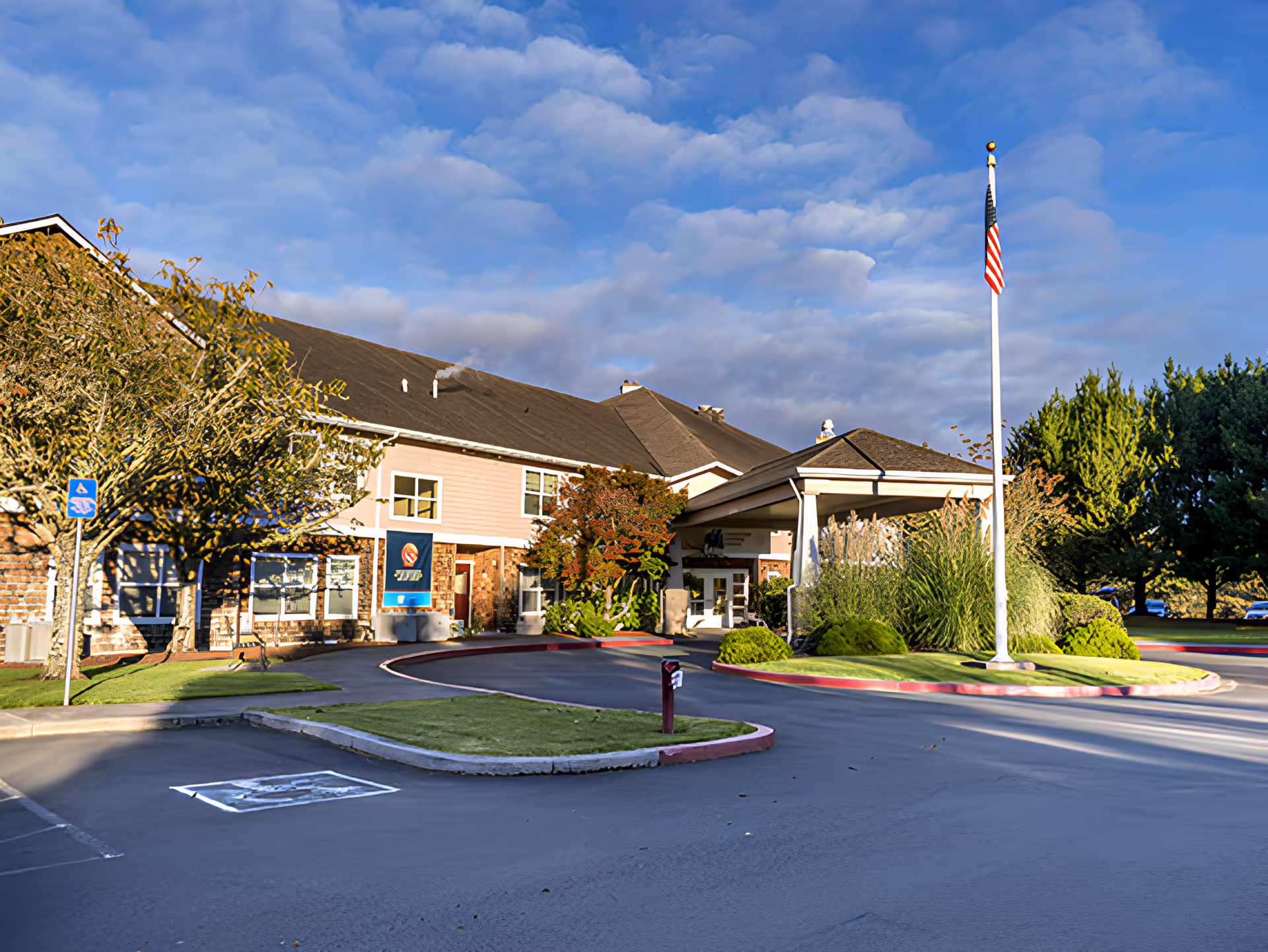 Exterior view of Avamere at Seaside senior living facility showing the main entrance with a covered drop-off area, an American flag on a flagpole, landscaped greenery, trees, and a clear blue sky with some clouds.