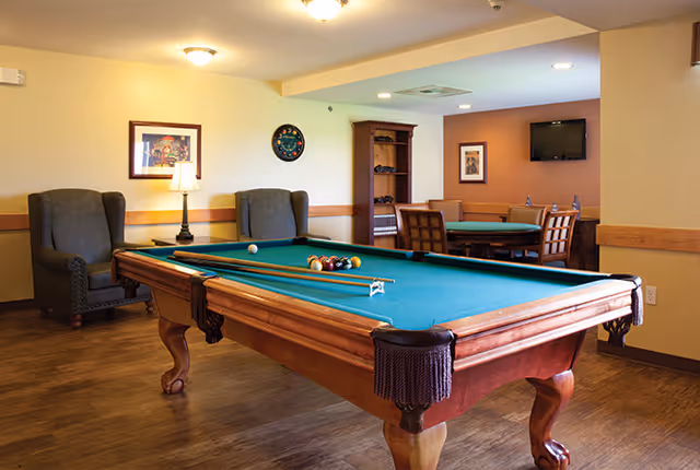 A recreational room featuring a pool table with cues and balls arranged on it, two upholstered armchairs against the wall, a round table with four chairs, a wall-mounted TV, framed artwork, and a clock on the wall.
