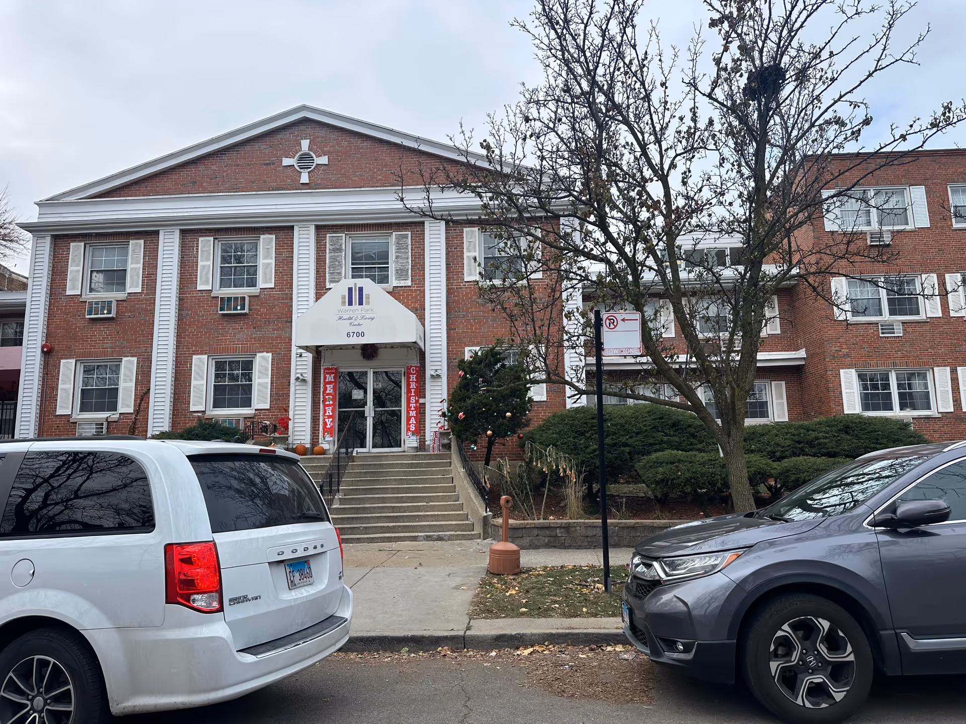 Brick multi-story senior living building with a central entrance and awning, front steps, shrubs, and parked cars along the street.