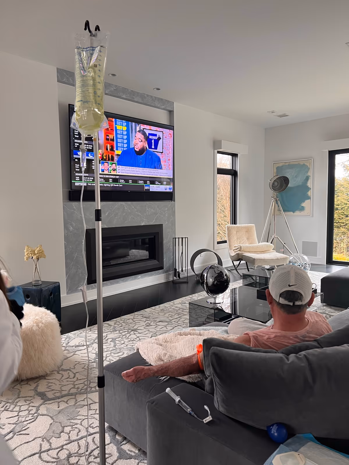 Resident sits on a gray sofa watching TV in a modern living room with IV stand