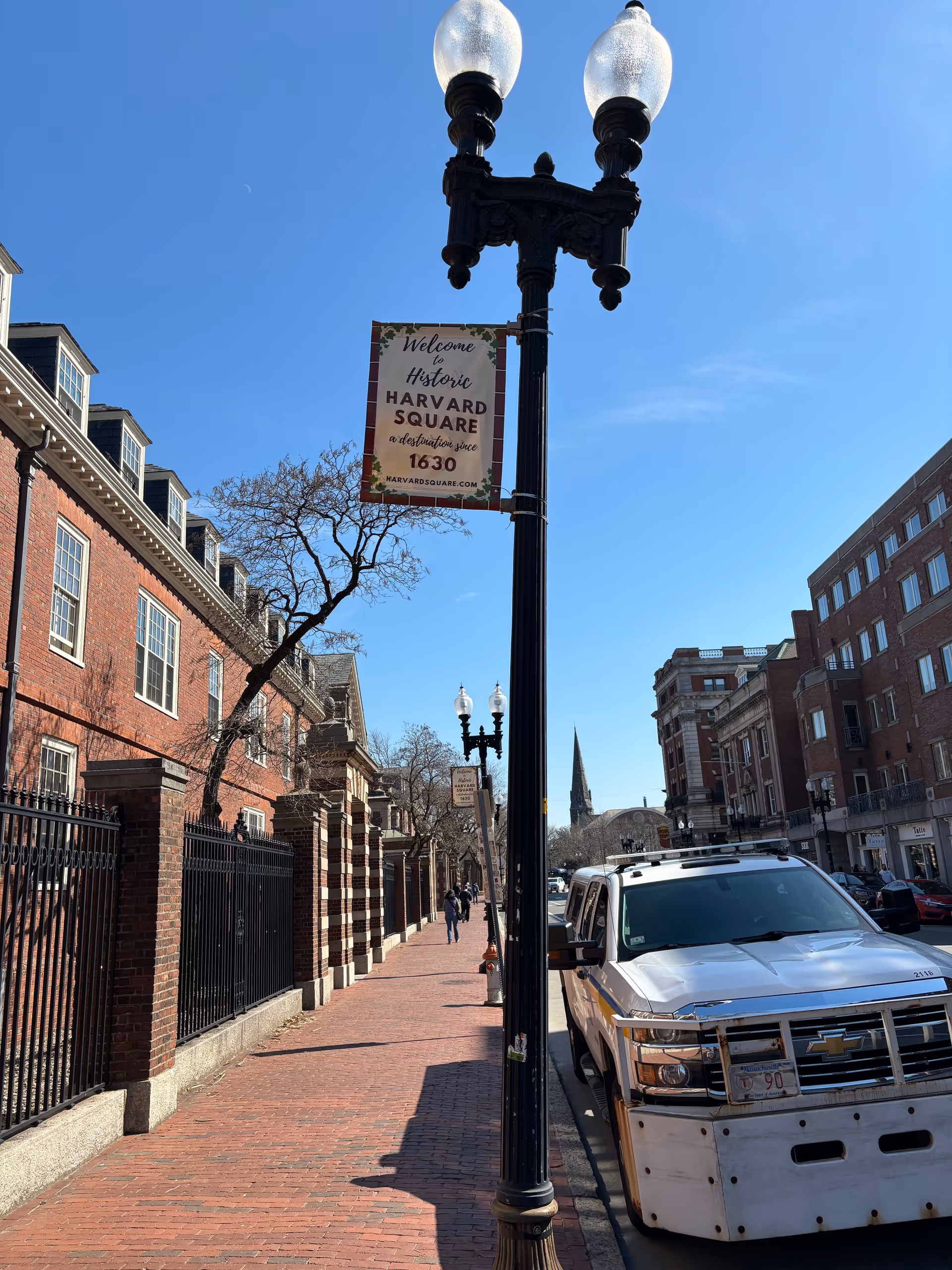 Sunny street scene in Harvard Square with brick buildings, a banner, and a parked white van.