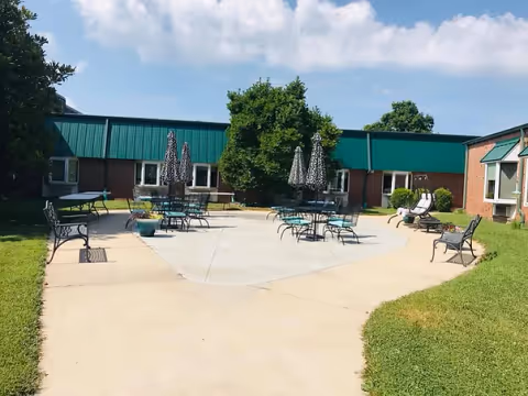 Courtyard patio with tables, chairs, umbrellas and benches surrounded by a single-story brick building under a partly cloudy sky.
