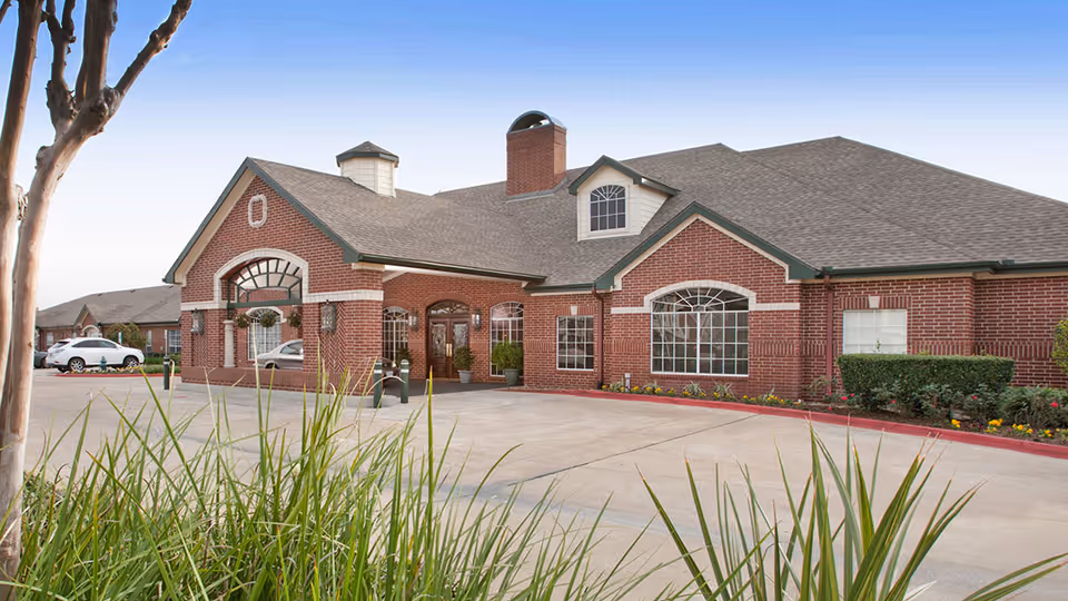 Exterior view of a single-story brick building with large windows and a peaked roof, surrounded by a paved driveway and landscaped with grass and shrubs.