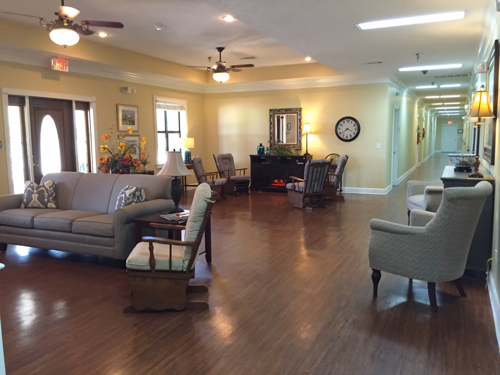 A spacious living room area with wooden flooring, featuring a gray sofa with patterned pillows, several cushioned wooden rocking chairs, and two upholstered armchairs. The room is decorated with lamps, a floral arrangement, a wall clock, and a mirror above a black electric fireplace. Ceiling fans and recessed lighting illuminate the space, and a long hallway extends from the living area.