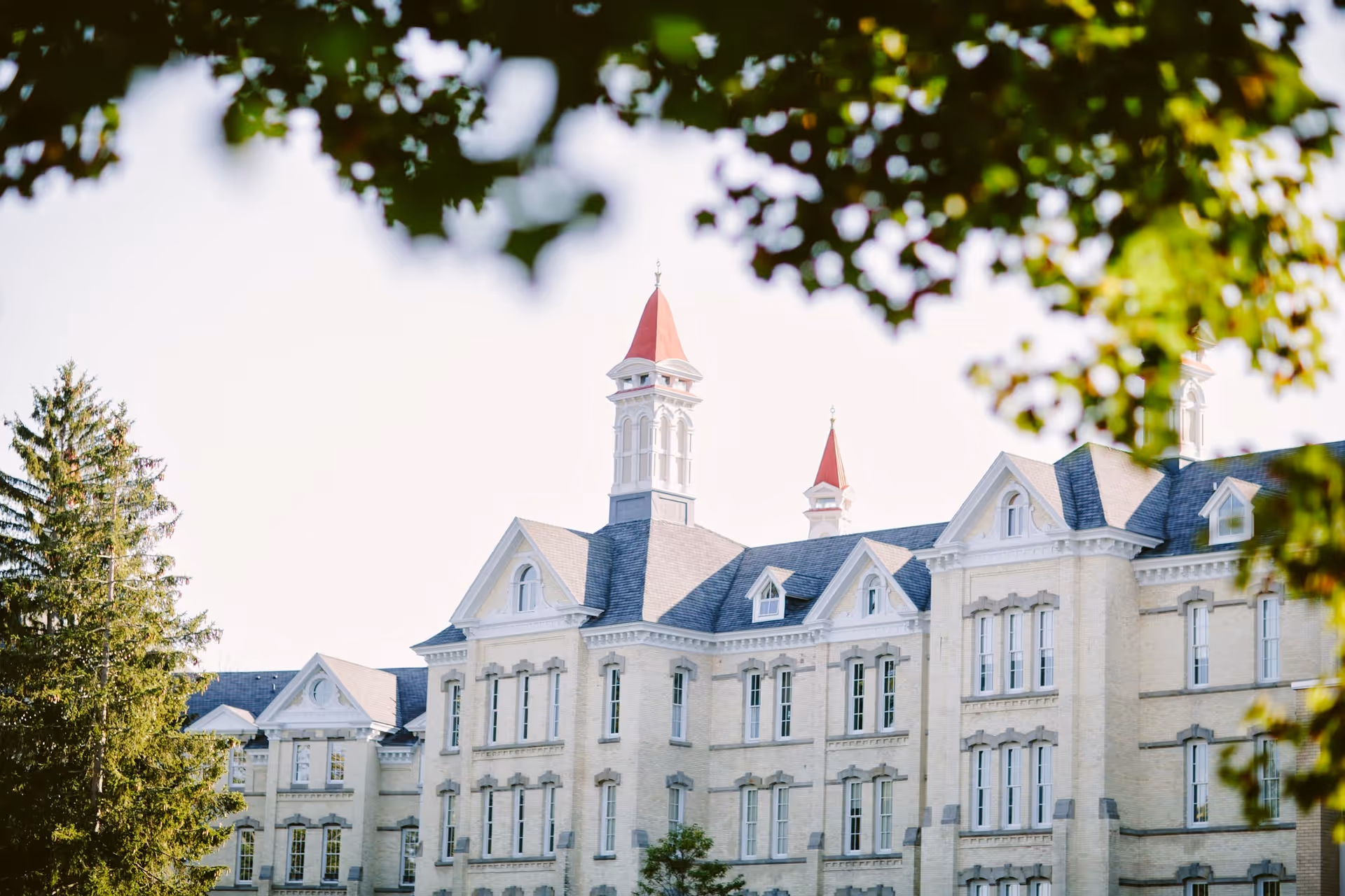 Historic multi-story brick building with red-roofed towers and dormer windows framed by leafy tree branches.