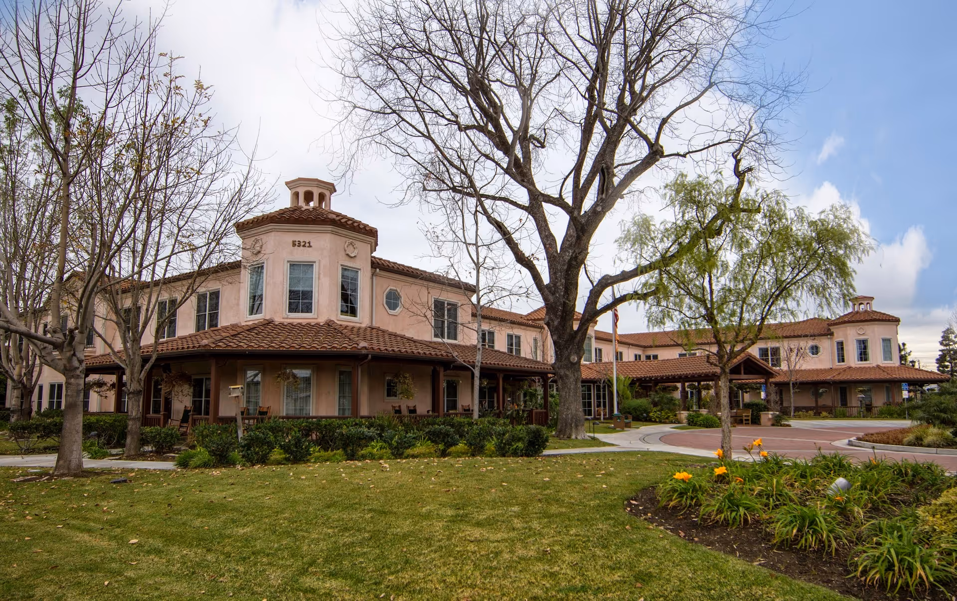 Exterior view of a two-story senior living facility building with a tiled roof, surrounded by trees and landscaped greenery under a partly cloudy sky.