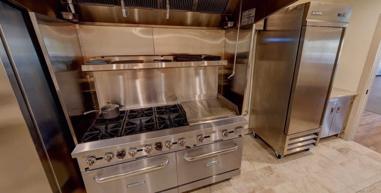 Commercial kitchen area with a large stainless steel stove featuring multiple burners and a griddle, a pot on one burner, and a stainless steel refrigerator next to it. The kitchen has a clean, professional look with stainless steel surfaces and tiled flooring.