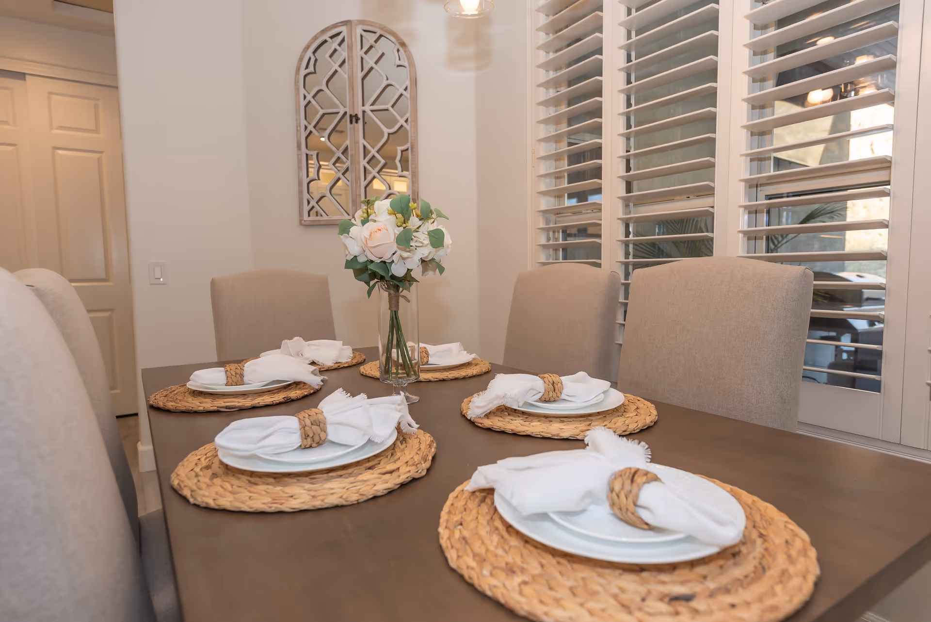 A dining table set for five with woven placemats, white plates, and white cloth napkins with woven napkin rings. A vase with white and pale pink flowers is placed in the center of the table. Beige upholstered chairs surround the table, and there are white plantation shutters on the windows in the background. A decorative wall mirror with a geometric pattern hangs on the wall.