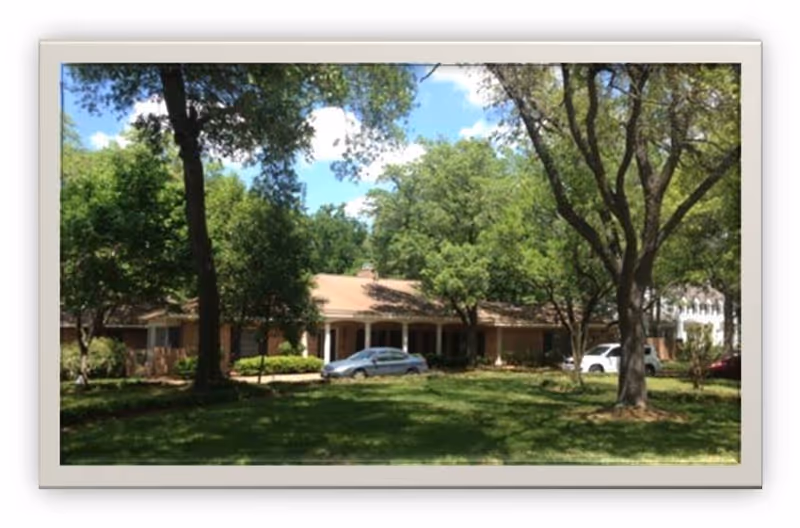 Single-story brick building with a covered entrance, cars parked in front and large trees on a grassy lawn.