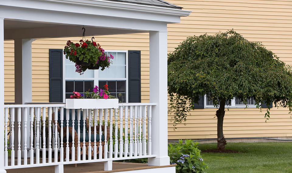 Front porch with white railing, a hanging basket and window box of flowers in front of a yellow-sided building and a small ornamental tree on the lawn.