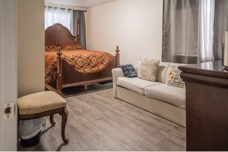 Furnished bedroom with a wooden four-poster bed, a beige sofa with pillows, an upholstered stool, and gray wood-look flooring.