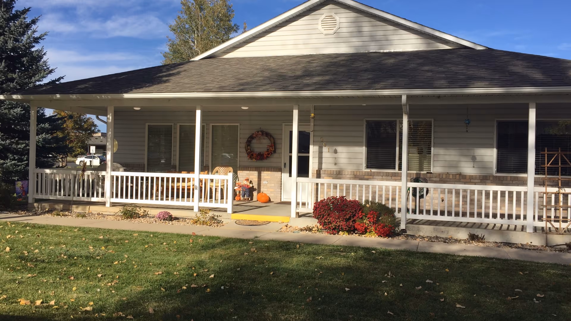 Front exterior view of a single-story building with a covered porch, white railing, and fall decorations including a wreath and pumpkins. There is a green lawn in the foreground and trees in the background under a blue sky.