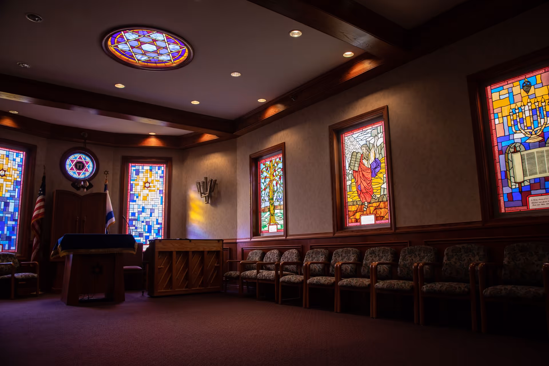 Interior of a room with stained glass windows depicting religious scenes, a row of cushioned chairs along the wall, a wooden podium with flags behind it, and a piano. The ceiling has recessed lighting and a circular stained glass light fixture.