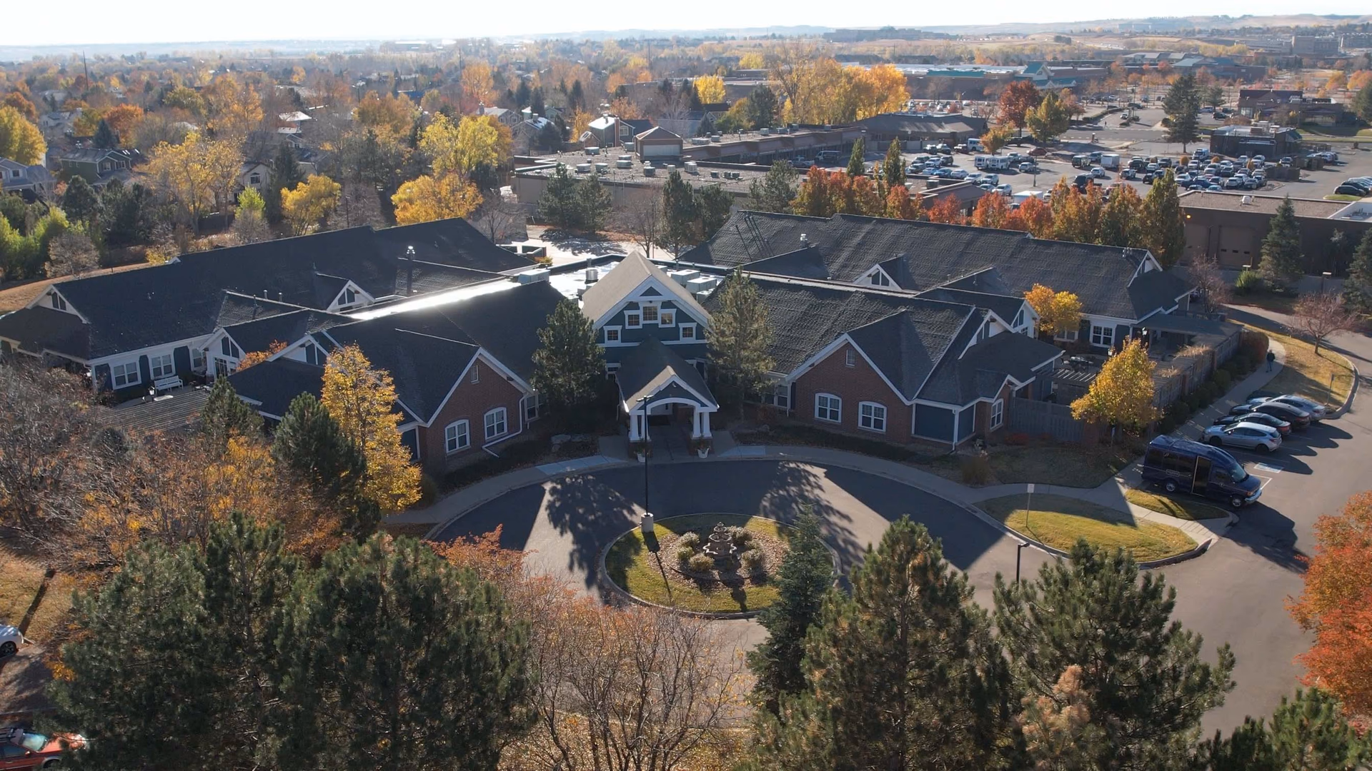 Aerial view of Balfour Cherrywood facility surrounded by trees with autumn foliage. The building has a circular driveway with a small landscaped island in the center. Several cars are parked along the driveway and in a parking lot nearby. The surrounding area includes residential homes and other buildings.