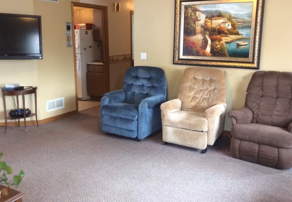 Three upholstered recliners in a communal living room with a wall-mounted TV, small table, and a framed seaside painting.