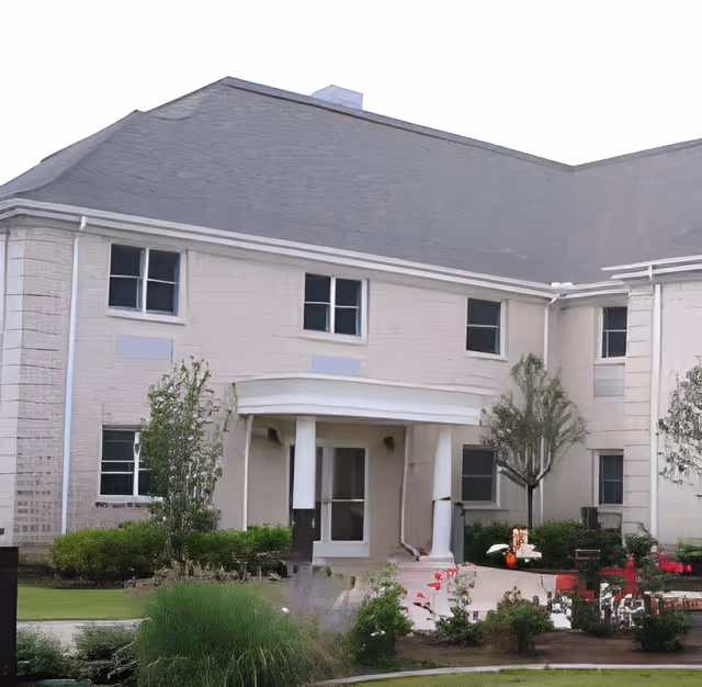 Front entrance of a two-story light-colored housing building with columns, windows, and landscaped shrubs.