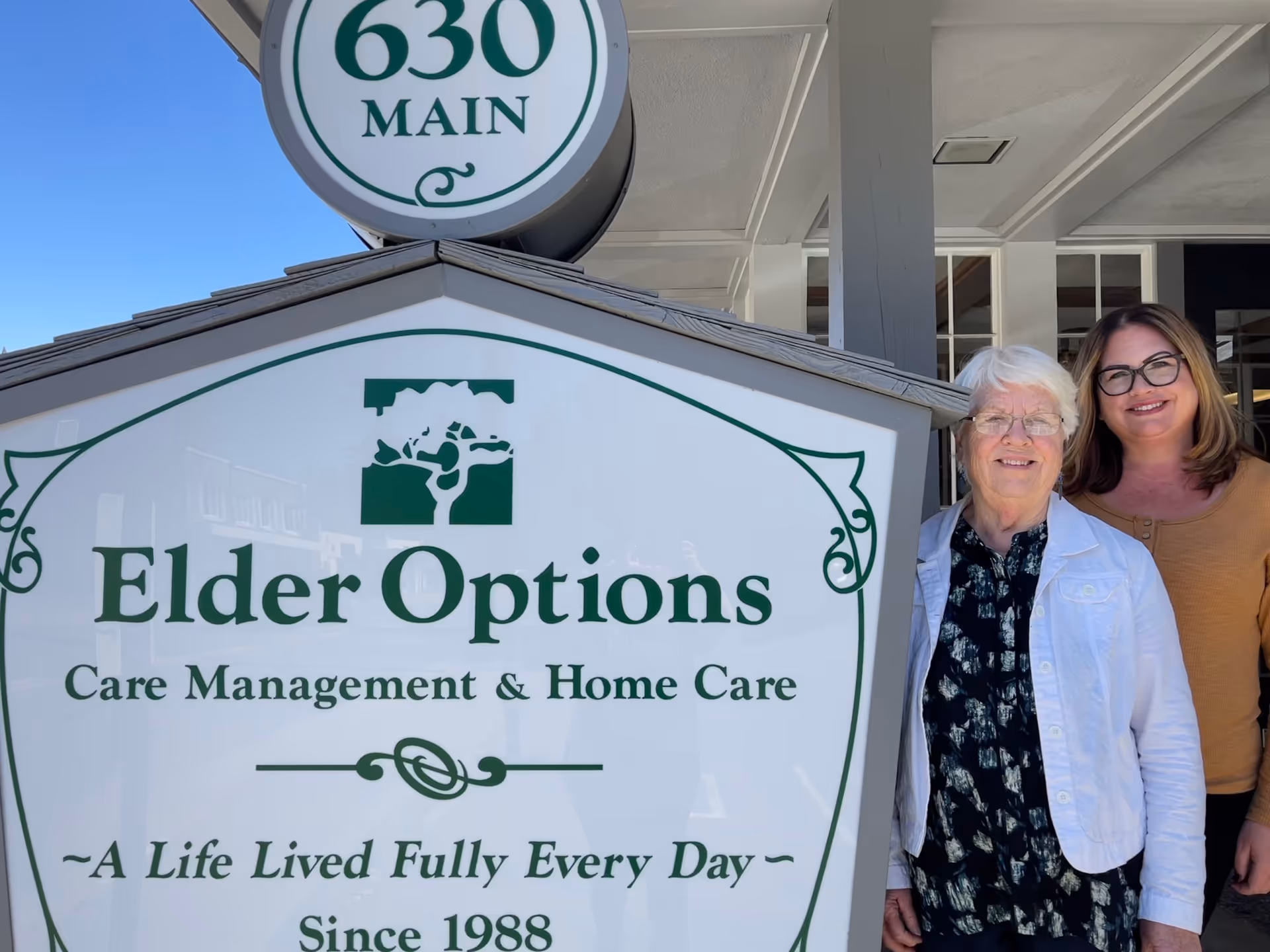 Two women stand beside an Elder Options sign outside a building entrance.