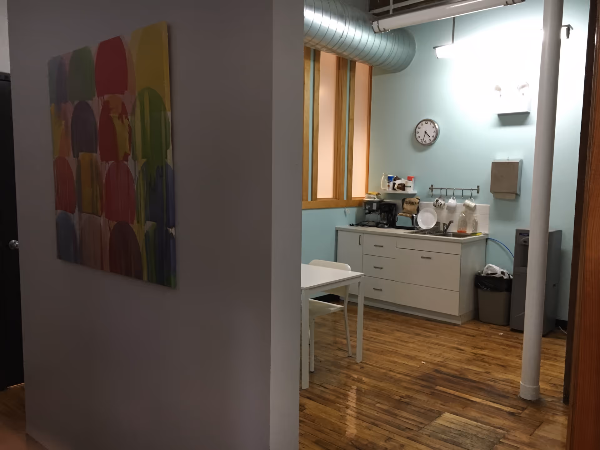 Small kitchen area with coffee machine, cabinets, table, and colorful wall art.