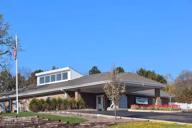 Single-story brick senior living facility with a covered entrance, flagpole, and landscaped front lawn under a clear blue sky.