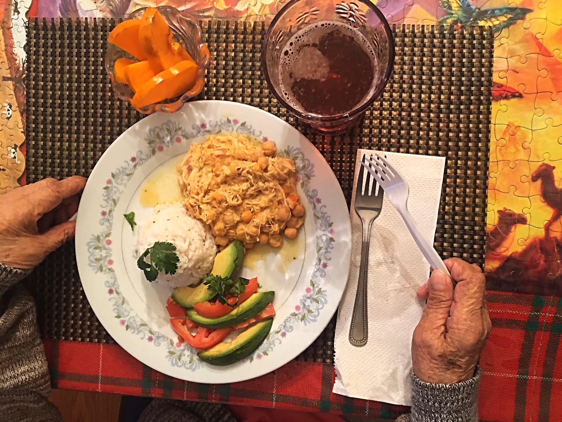 Top-down view of a person's hands at a table with a plate of rice, chickpea stew, avocado and tomato slices, a glass of drink, and a small bowl of orange slices.