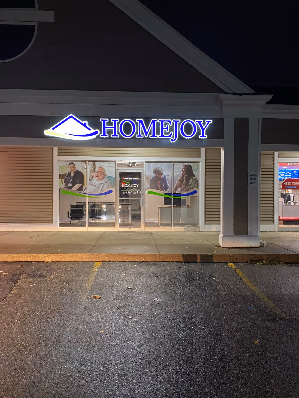 Nighttime exterior of a HomeJoy storefront with illuminated sign, glass doors, and resident posters.