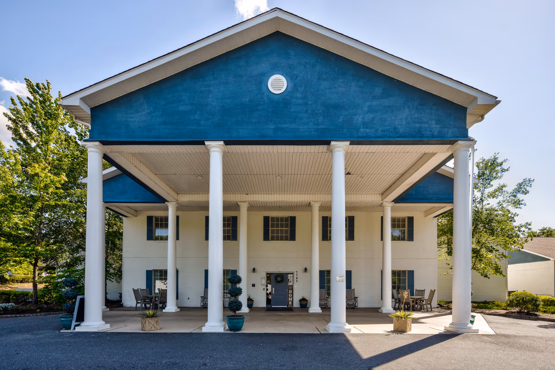 Front exterior view of a two-story building with white columns supporting a blue triangular pediment. There are rocking chairs and potted plants on the covered porch area, with trees and clear sky in the background.