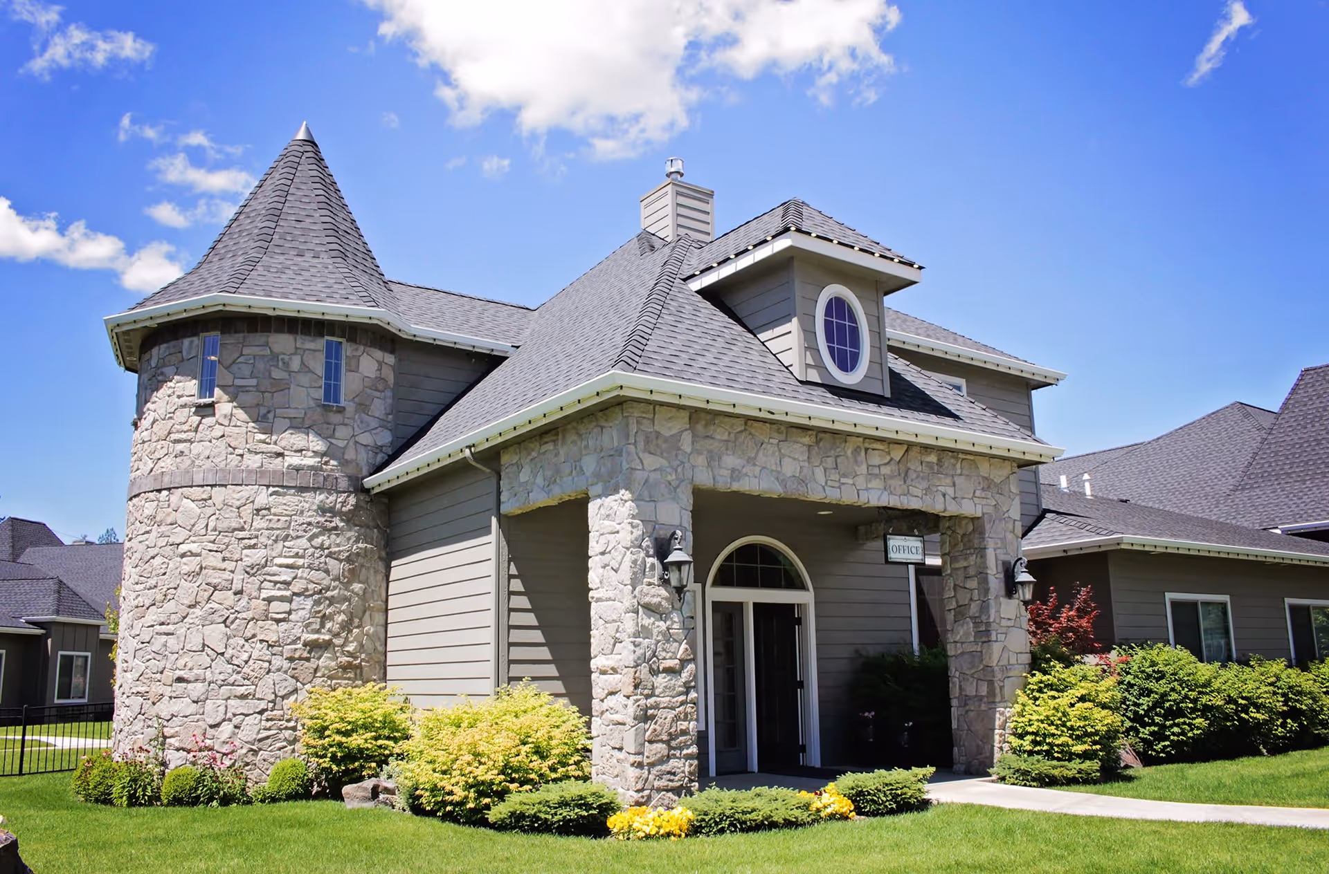 Stone-and-siding senior living building with a turret and a landscaped entrance labeled "Office" under a blue sky.