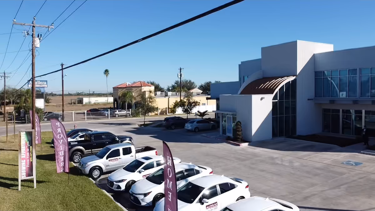 Modern white building with glass facade and curved awning, parking lot and purple flags outside.