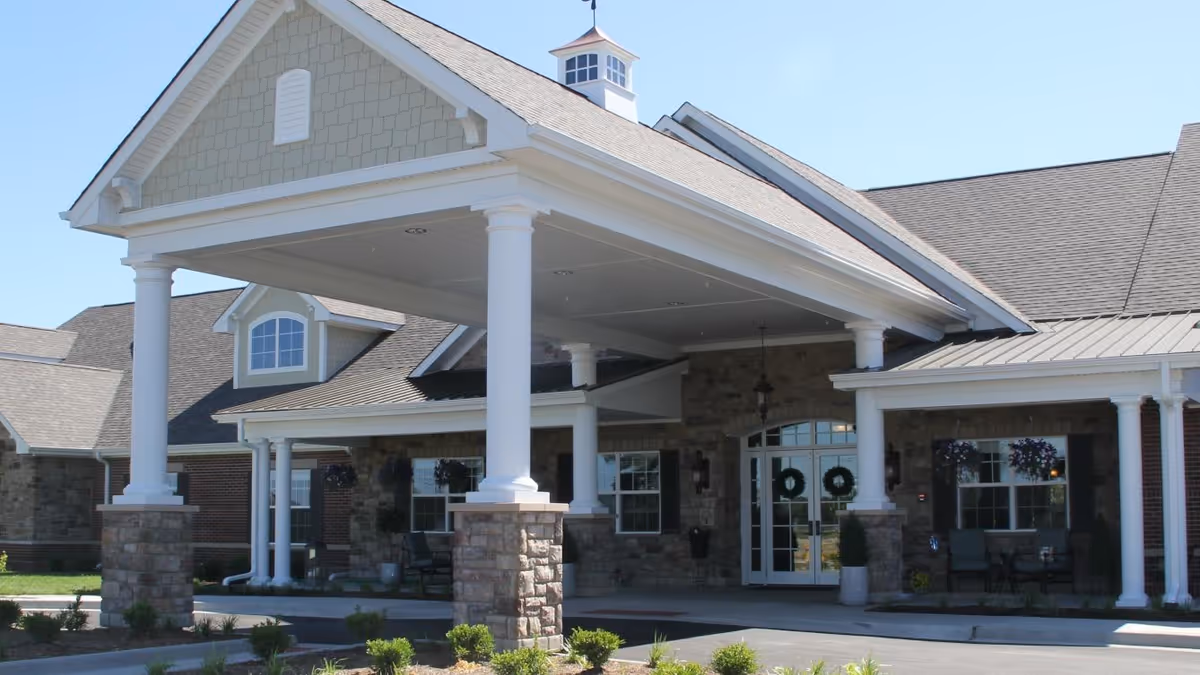Front exterior view of a senior living facility with a covered entrance supported by white columns on stone bases, a peaked roof with a cupola, and a seating area with chairs and hanging flower baskets near the entrance.