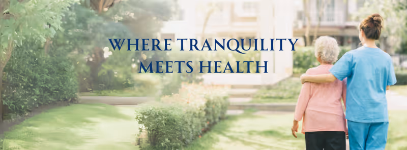 Caregiver and elderly woman walk outdoors along a landscaped path at a serene facility.