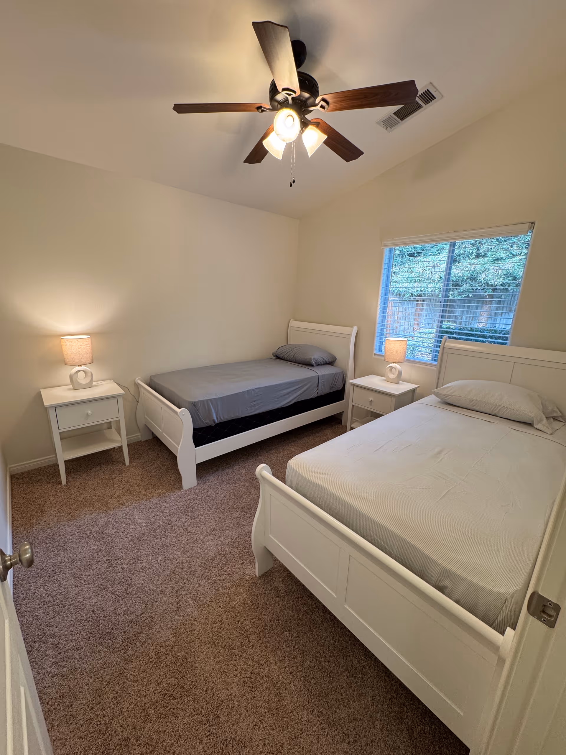 Two white twin beds with matching nightstands and ceiling fan in a neutral bedroom.