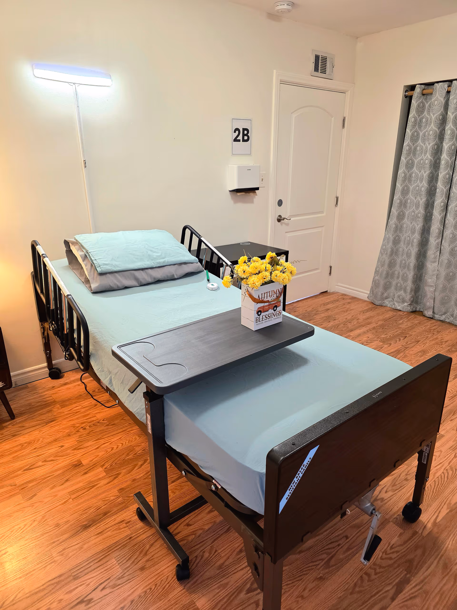 A hospital-style bed in a neat patient bedroom with a tray and flowers.