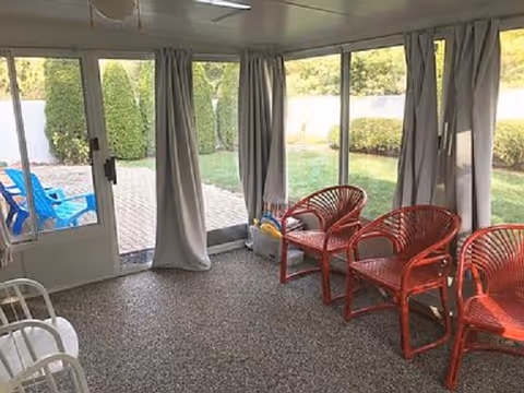 Enclosed sunroom with red wicker chairs, white curtains, and glass doors to a garden.