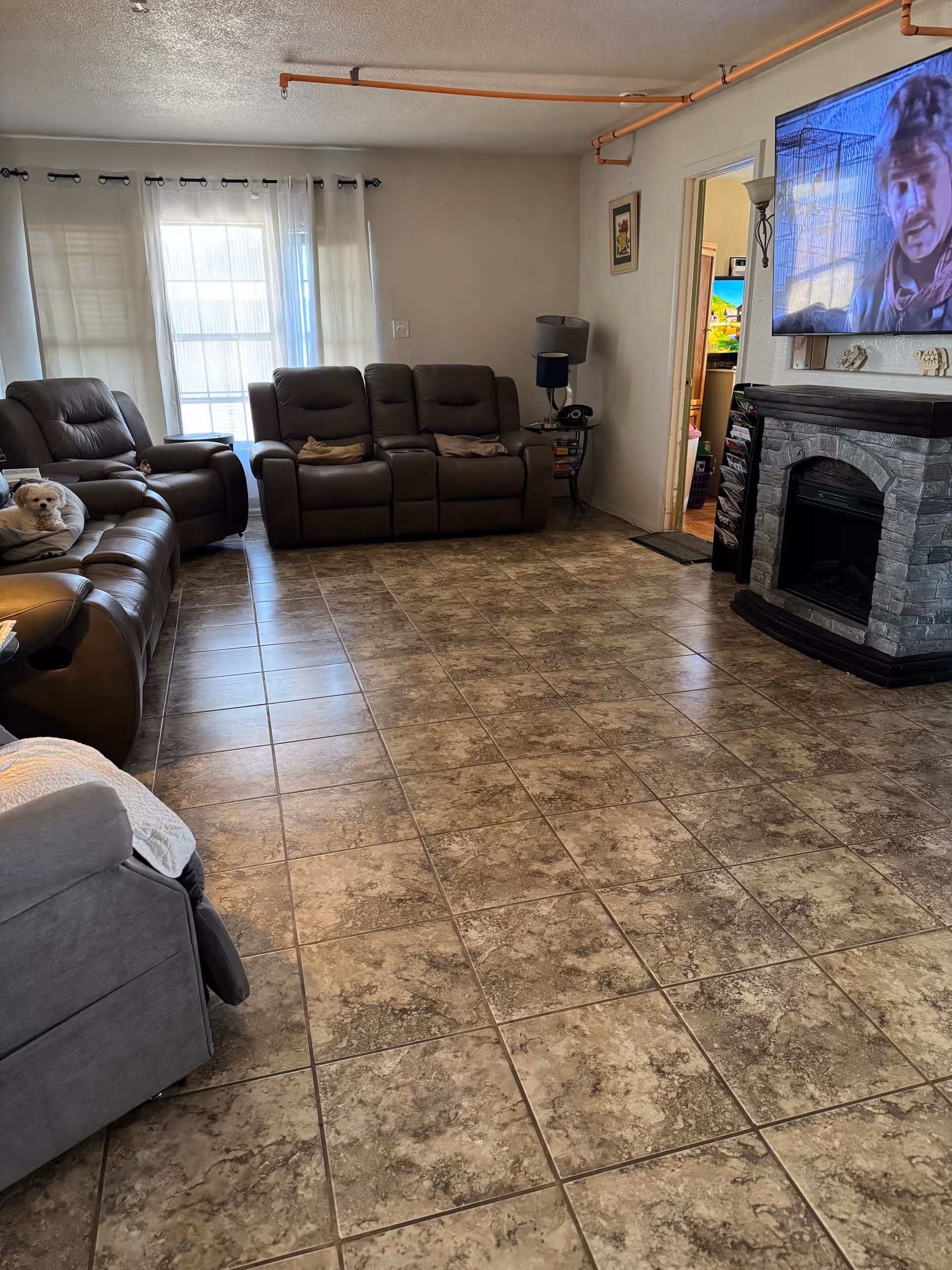 Spacious living room with tile floor, brown reclining sofas, a wall-mounted TV above a faux-stone fireplace, and a small dog on a couch.