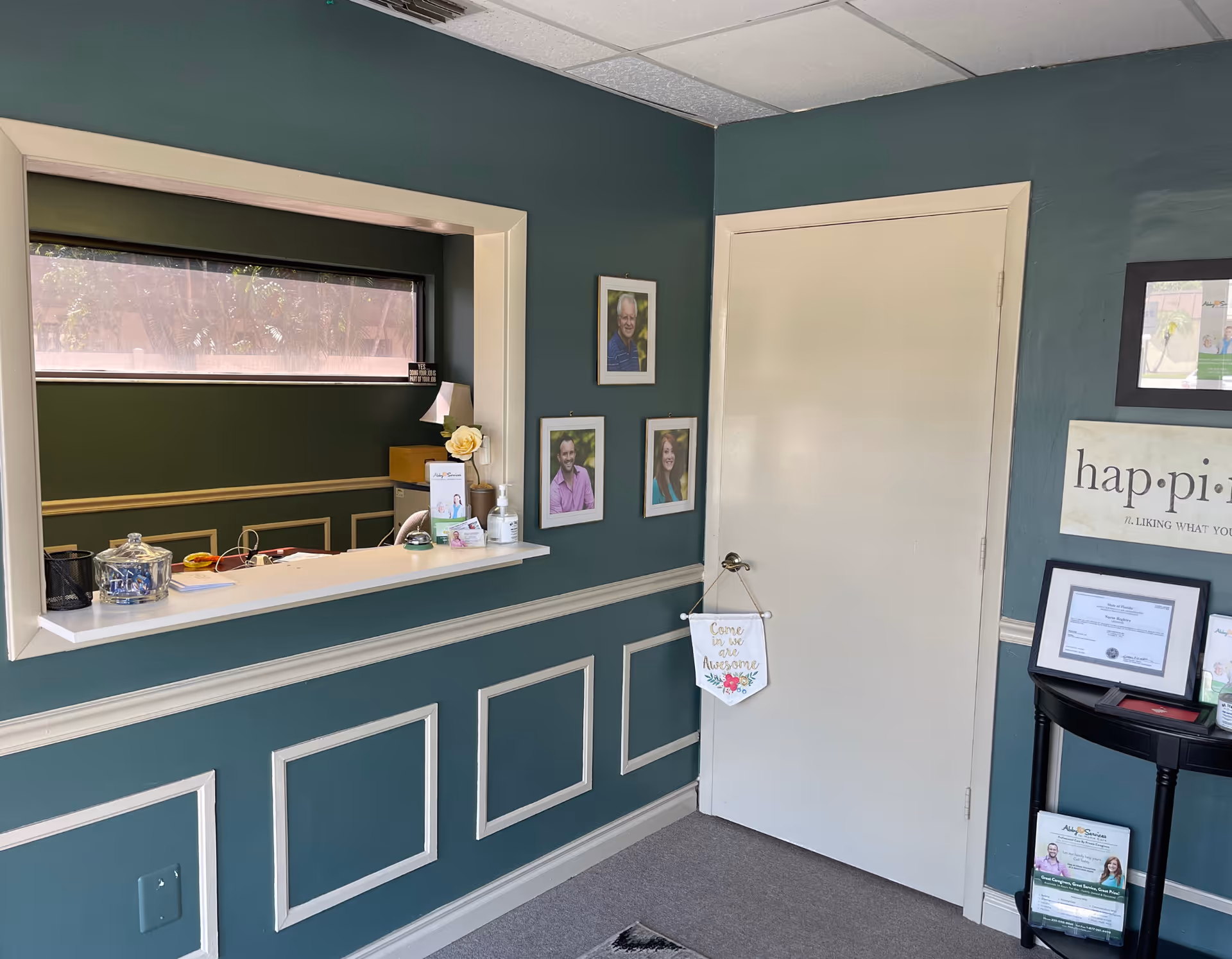 Teal-walled reception area with a window counter, framed photos, and a white door.