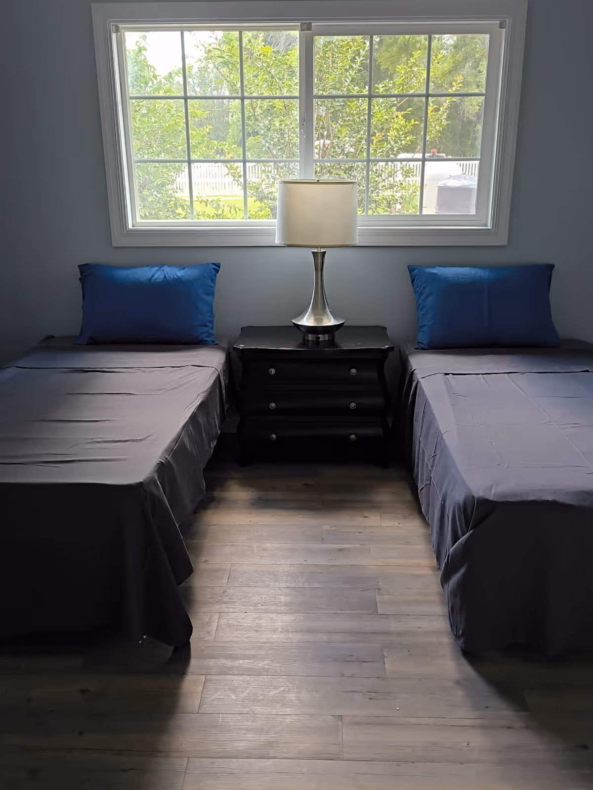 Two twin beds with blue pillows flank a nightstand and lamp beneath a large window.
