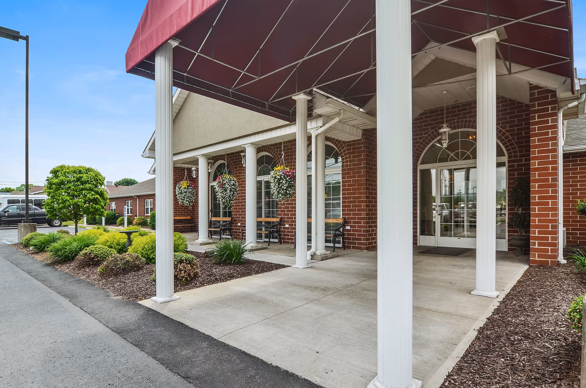 Brick facility entrance with white columns, maroon canopy, and hanging flower baskets.