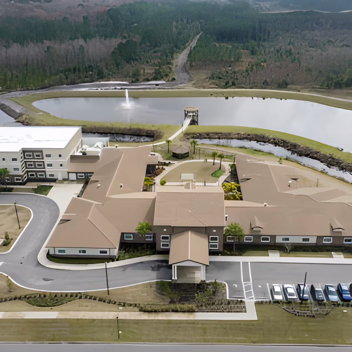 Aerial view of Discovery Village At Deerwood Assisted Living & Memory Care facility showing a large building complex with beige roofs, surrounding parking lot with cars, landscaped grounds, a pond with a fountain, and a wooded area in the background.