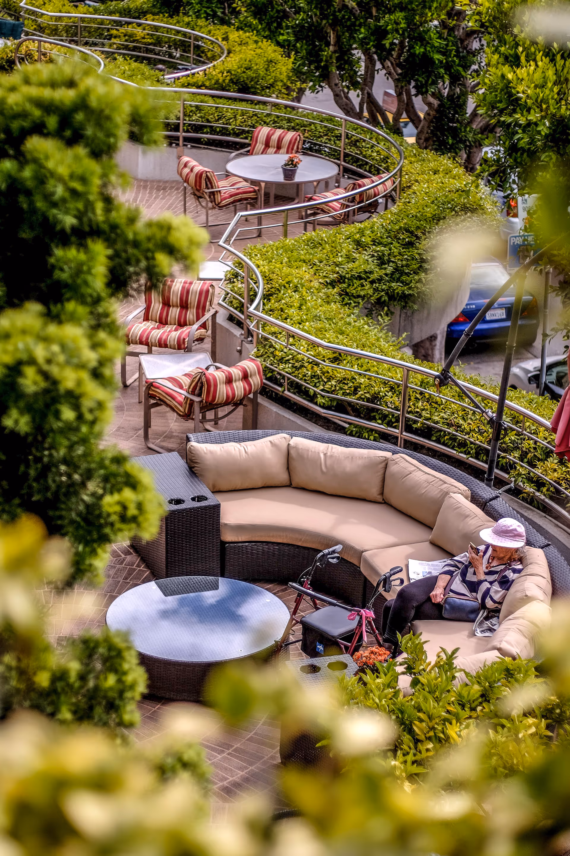 Outdoor curved patio with cushioned wicker seating, round tables, striped chairs, lush hedges, and an elderly woman sitting with a walker.