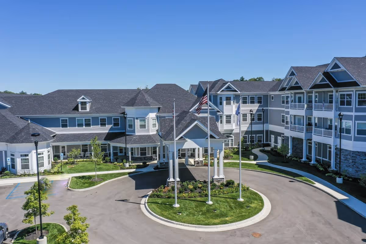 Exterior view of a large, modern senior living facility with multiple connected buildings, a circular driveway with a covered entrance, three flagpoles with flags, well-maintained landscaping, and clear blue sky.