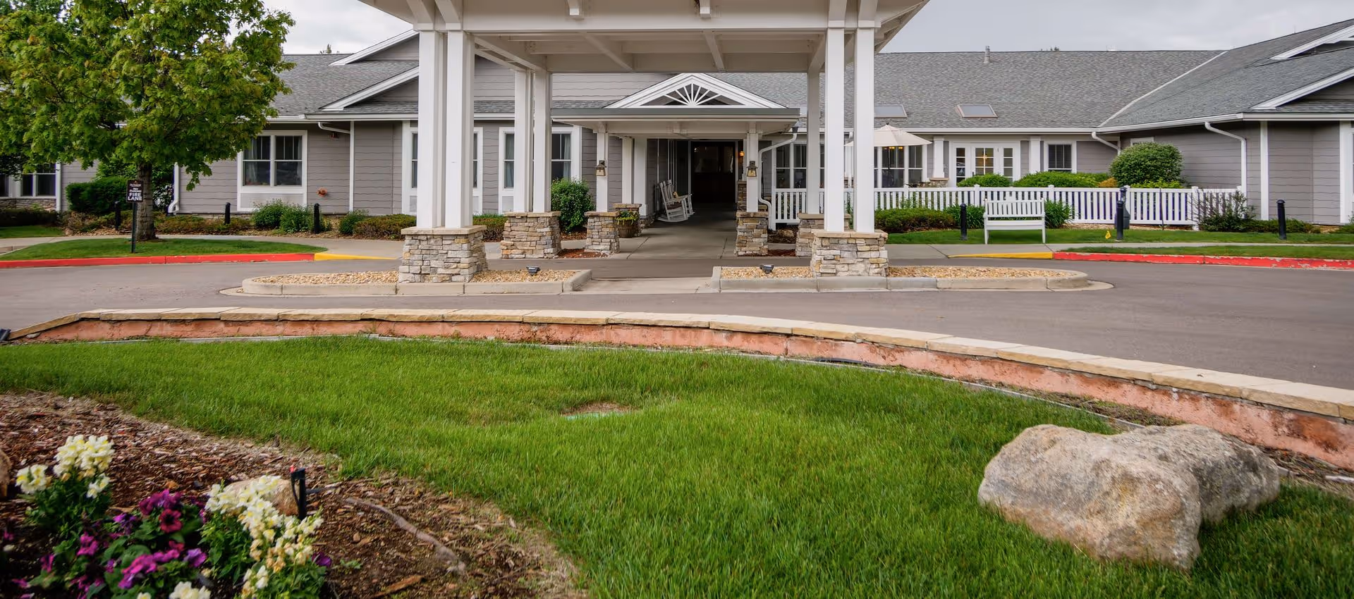 Covered main entrance/porte-cochere of a low-rise senior living building with stone pillars, white benches, and landscaped lawn in front.