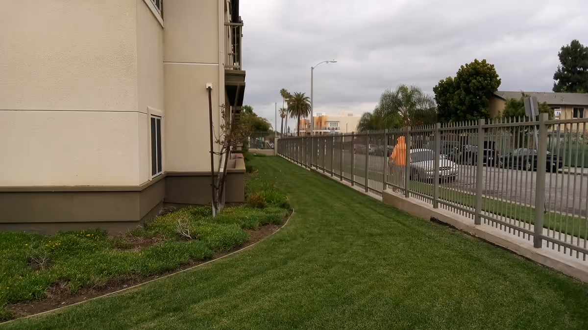 Exterior view of a beige senior housing building with a grassy side lawn and metal fence.