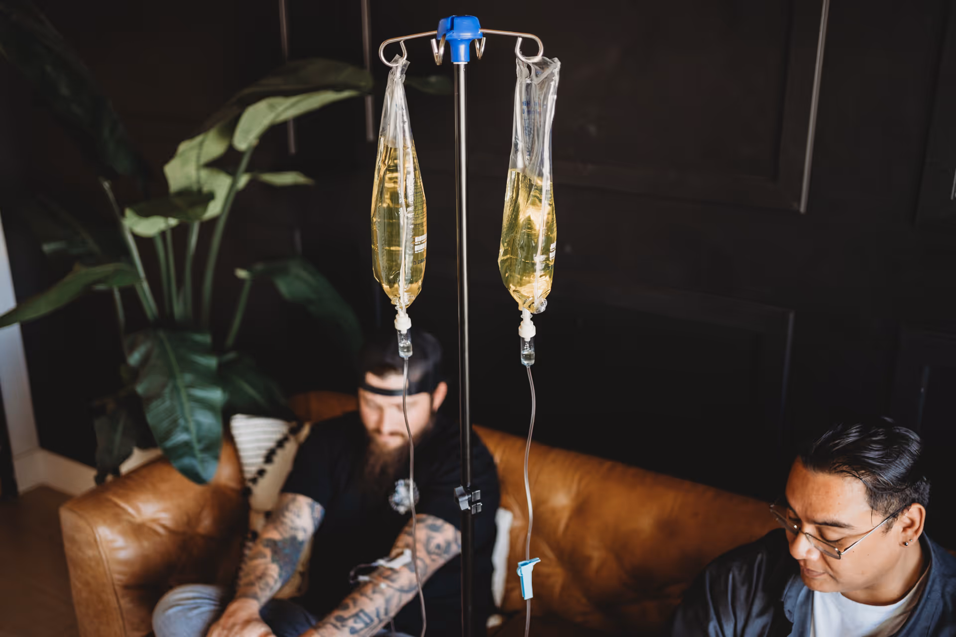 Two men sit on a couch indoors with IV drip bags in the foreground.